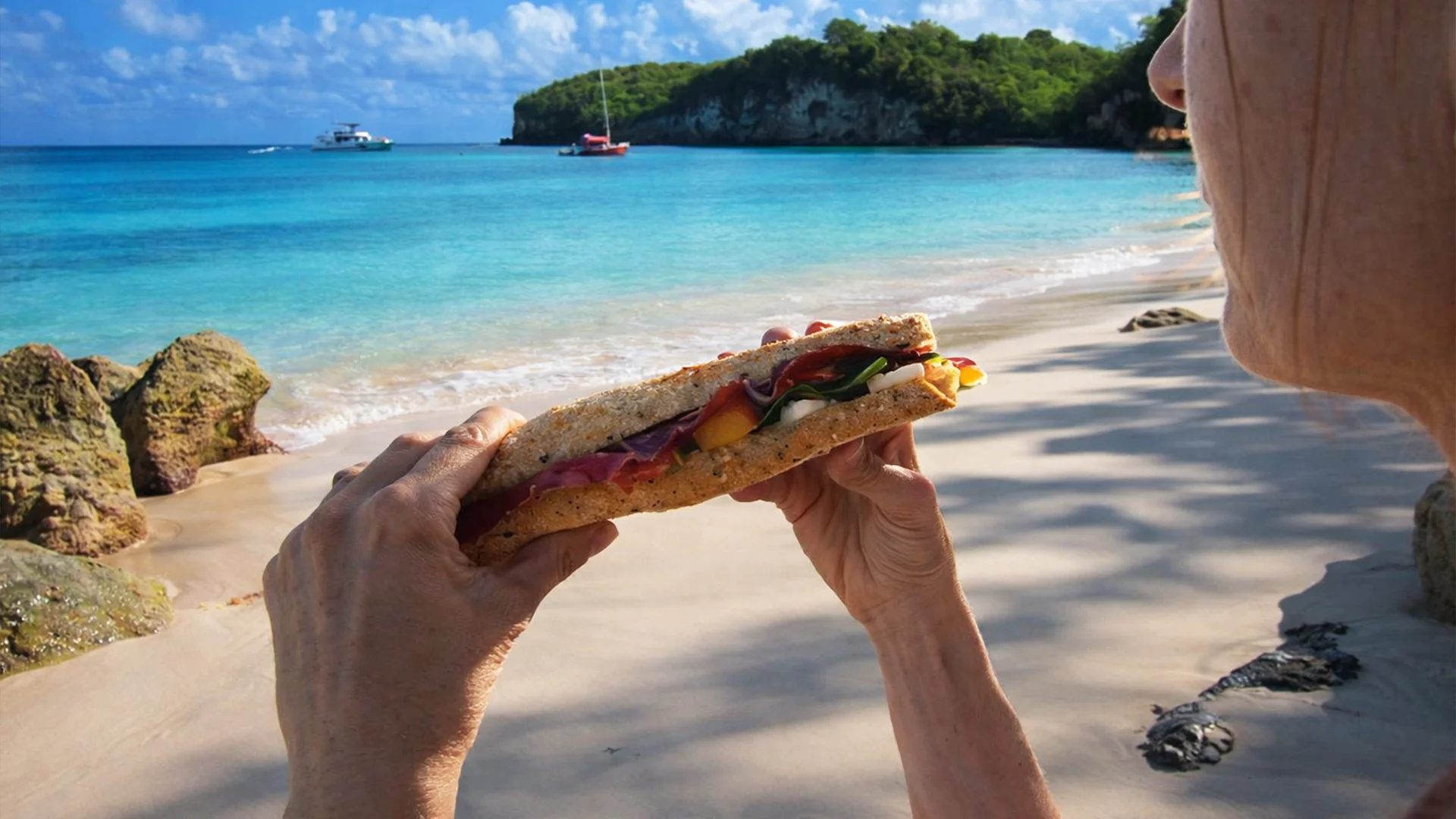 Pique-nique sur une plage sauvage de Marie-Galante en Guadeloupe, sandwich à la main face à une eau turquoise lors d’une excursion en bateau.