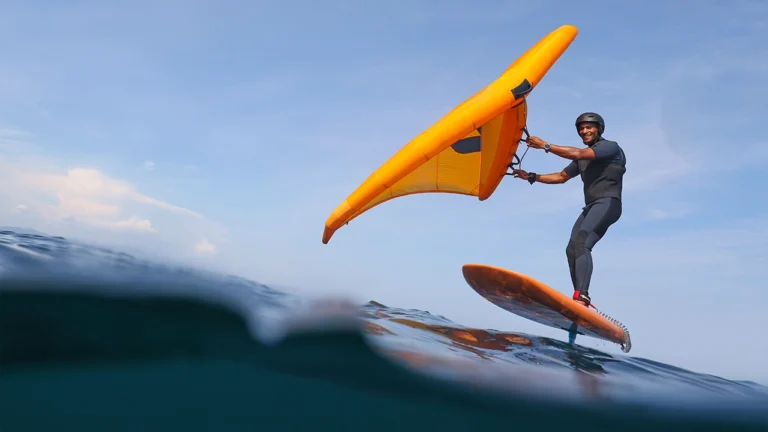 Homme pratiquant le wingfoil avec une aile orange sur une planche à foil au-dessus de l’eau.