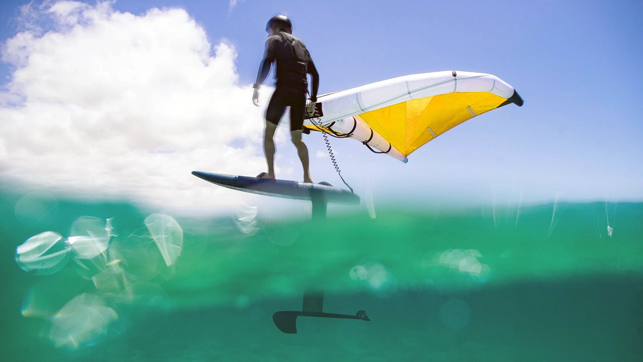Pratiquant de wingfoil en mer turquoise avec une aile jaune et blanche, planant au-dessus de l’eau.